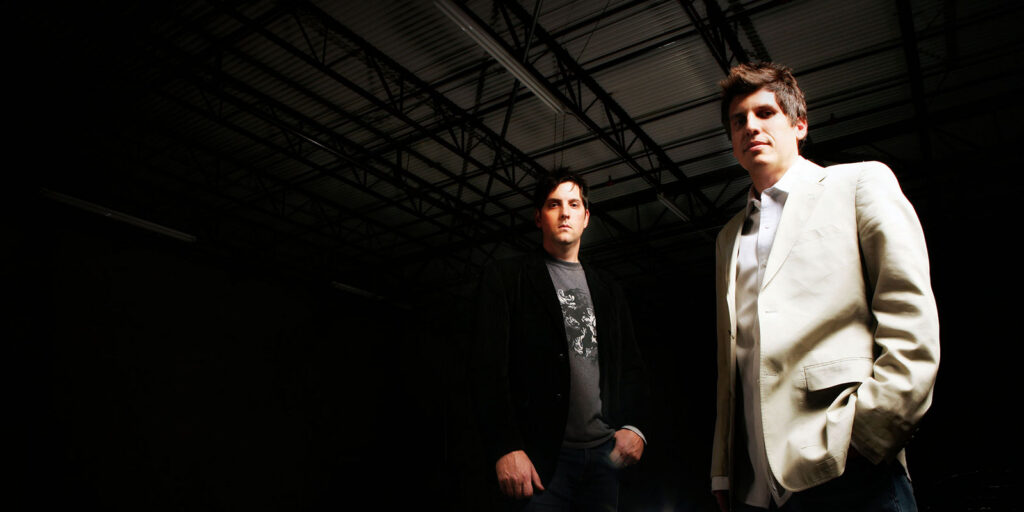 Two men stand indoors under industrial ceiling beams; one wears a dark jacket and T-shirt, the other a light-colored suit jacket and shirt, discussing brand evolution for Designing Ardent.