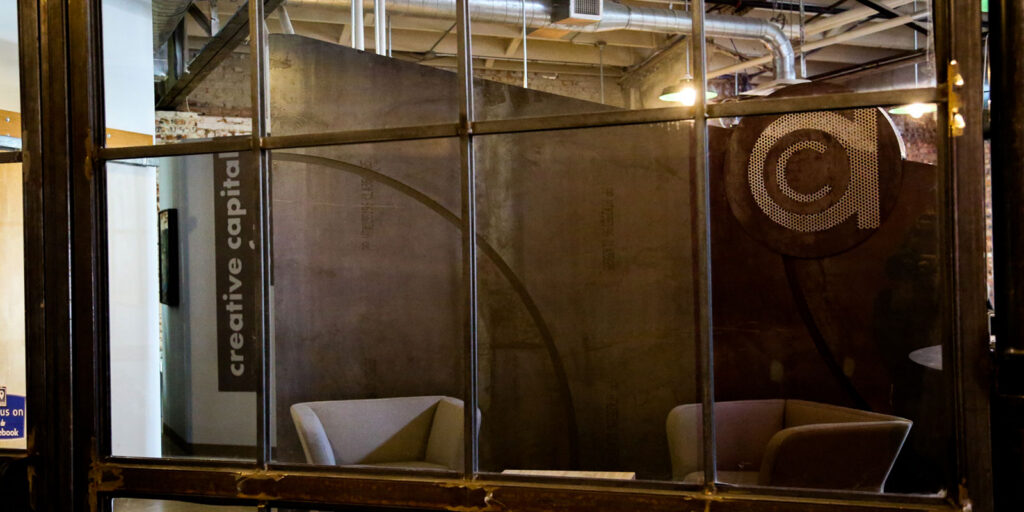 View through a glass window of a modern office space with two gray chairs, a small table, and a wall displaying the words "creative capital," reflecting Ardent’s commitment to brand evolution.